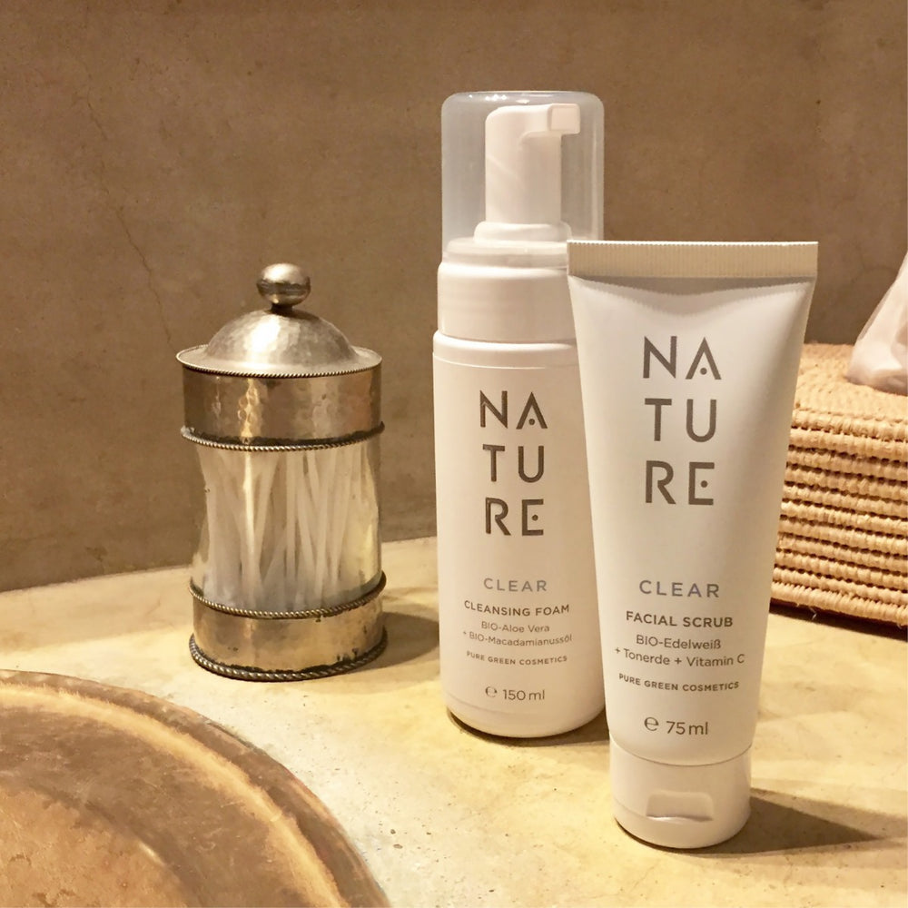 Reinigungsschaum mit Aloe-Vera & Macadamianussöl auf Badezimmerablage, neben Peeling und Glasdose mit Wattestäbchen. Bio & vegan, für gründliche Reinigung und gleichmäßiges Hautbild.
