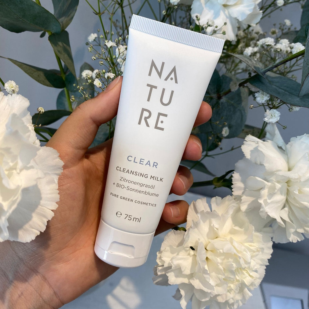 Hand hält 75 ml Tube NATURE CLEAR Reinigungsemulsion mit BIO-Sonnenblumenöl und Zitronengras, umgeben von weißen Blumen und Eukalyptus, für sanfte, erfrischende Hautpflege.
