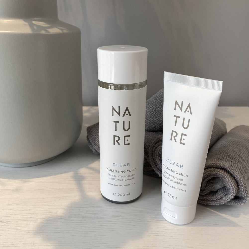Reinigungstonic mit Rotklee-Extrakt & Aloe-Vera für gründliche Reinigung & verfeinerte Poren, neben Cleansing Milk, grauem Handtuch und Keramiktopf auf Holzoberfläche.