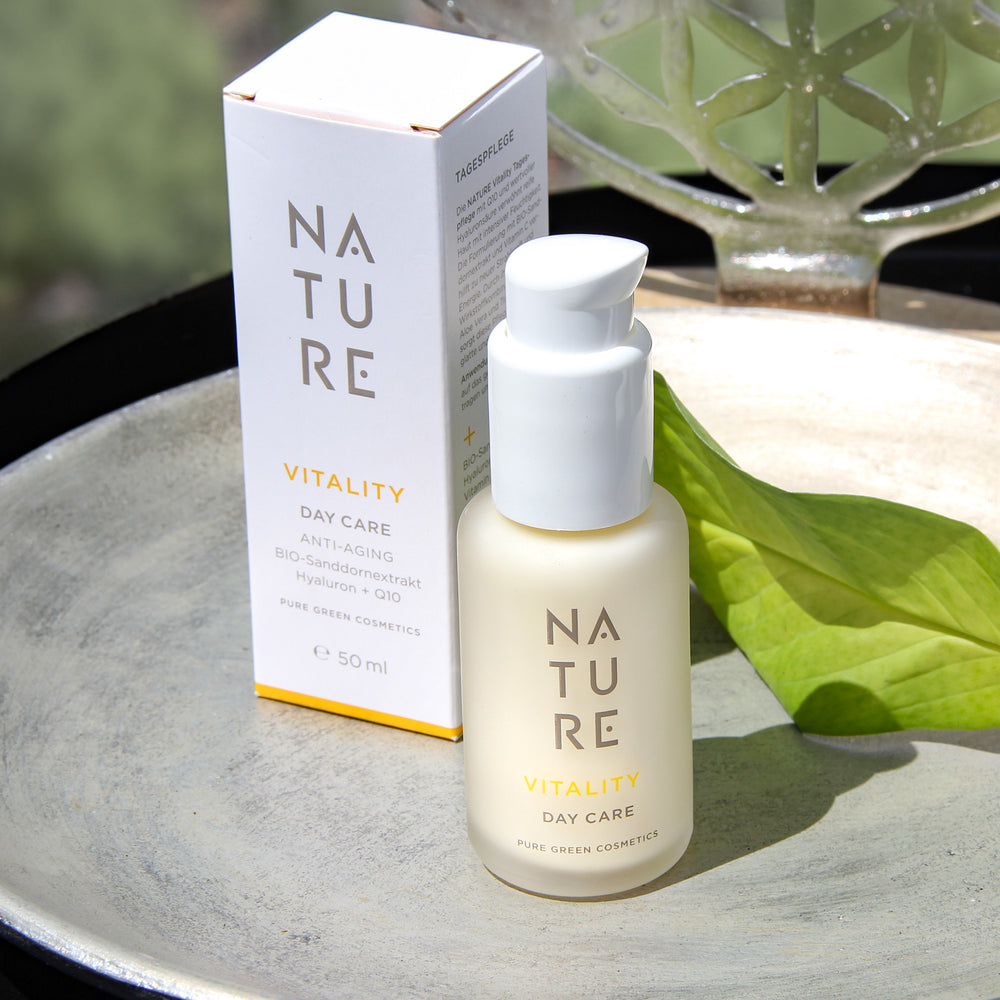Vitality Tagescreme mit Q10 & Sanddornextrakt, im mattierten Glasflakon mit Pumpverschluss, auf flacher Schale neben Blatt, bietet strahlende, samtige Haut. Bio & vegan.