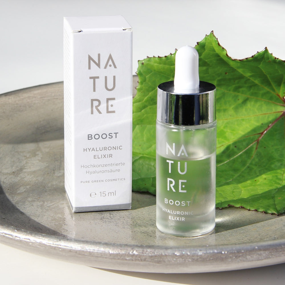 Kleine Tropfflasche „NATURE BOOST Hyaluronic Elixir“ und Schachtel auf Metallschale, mit grünem Blatt im Hintergrund; enthält hochkonzentrierten Hyaluron-Komplex für Feuchtigkeit und Elastizität.
