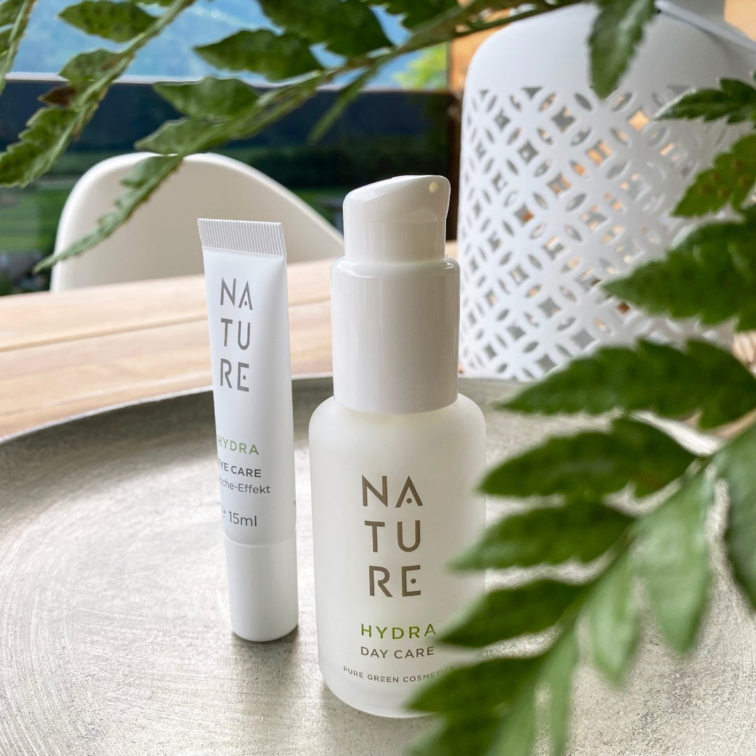 Zwei NATURE HYDRA Produkte: links HYDRA Eye Care Tube, rechts Tagescreme mit Gurkenwasser & Hyaluronsäure Pumpflasche, dekoriert mit Grün auf Metalltablett im Freien.
