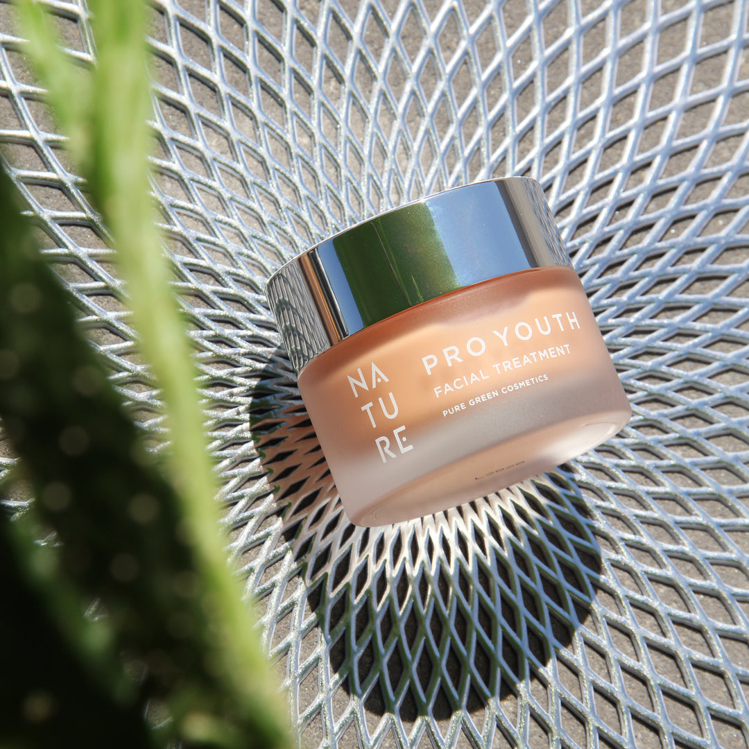 Pro-Youth Gesichtscreme mit Astaxanthin & Hyaluronsäure in einem mattierten Glasbehälter auf Metallgitter, umgeben von unscharfen grünen Blättern, für Anti-Aging-Pflege.