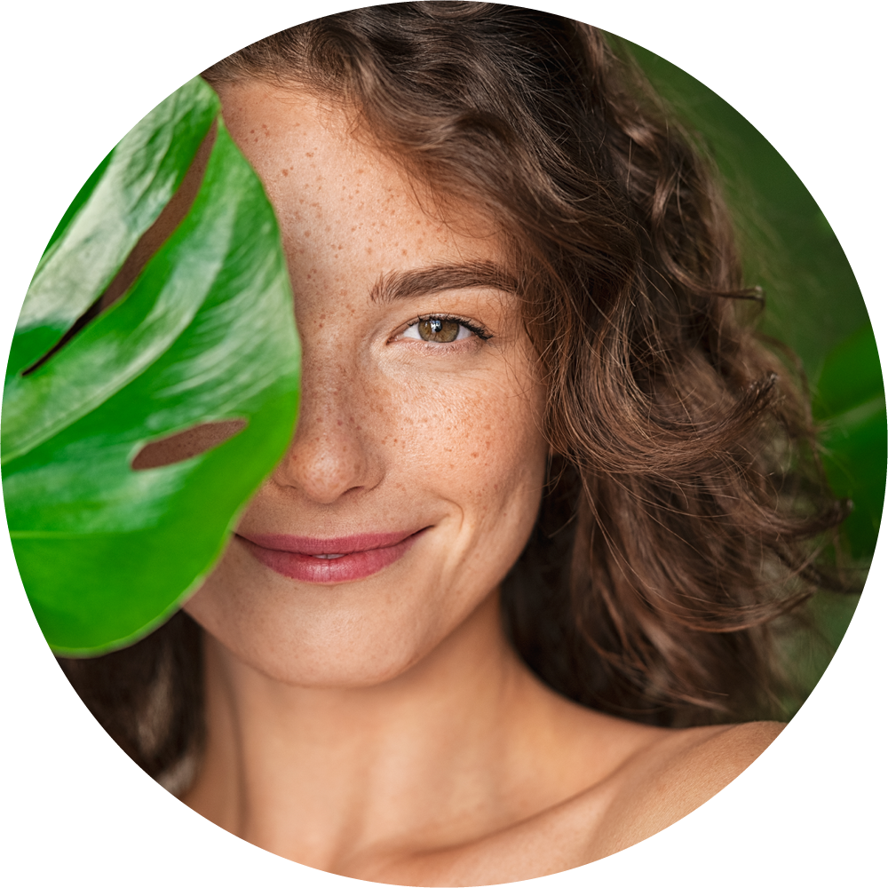 Porträt einer jungen Frau mit Sommersprossen und welligem braunem Haar, teilweise von einem großen, grünen Monstera-Blatt verdeckt. Frischer, natürlicher Look.