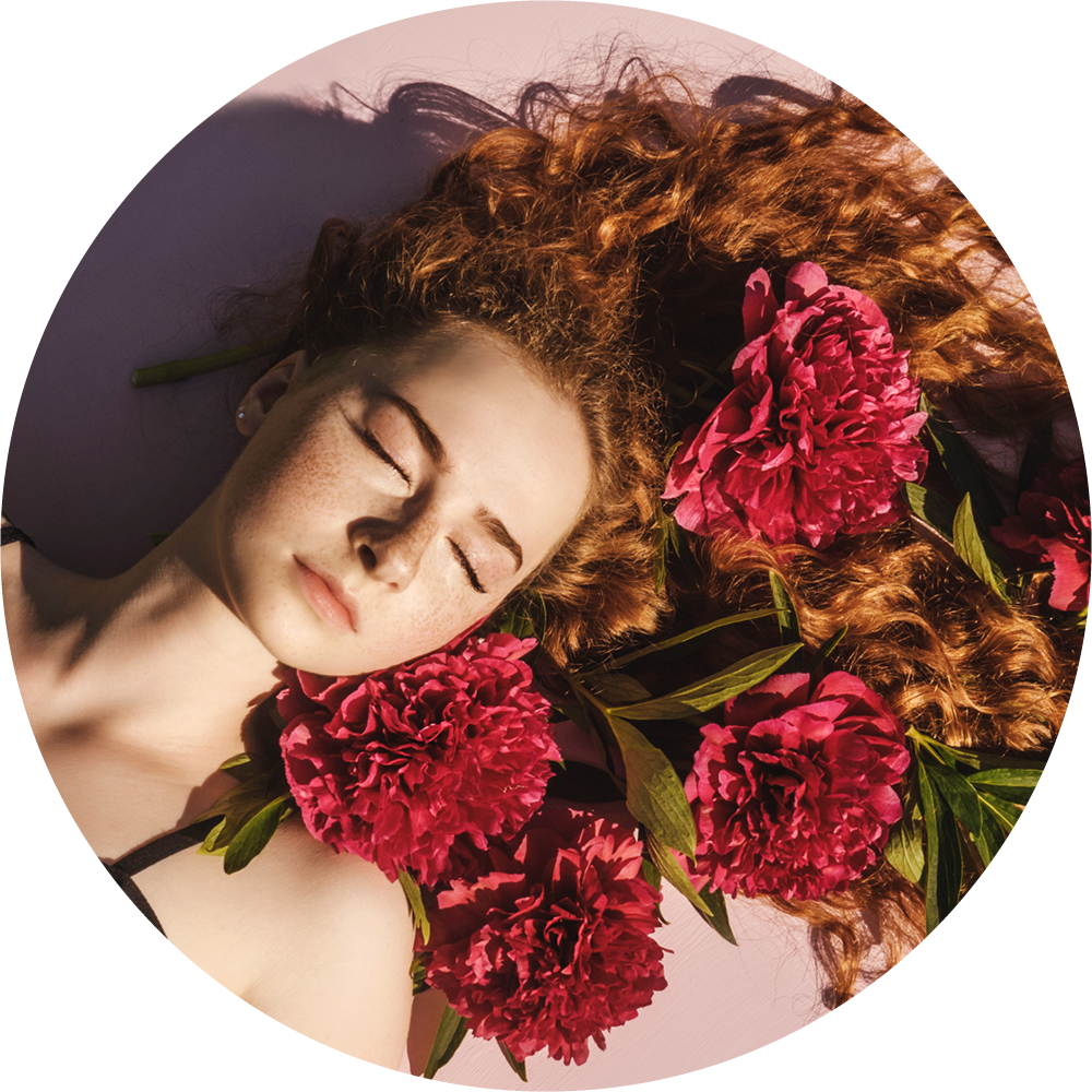 Eine junge Frau mit Sommersprossen und langen, roten Locken liegt mit geschlossenen Augen, umgeben von großen, rosa Pfingstrosenblüten und grünen Blättern.