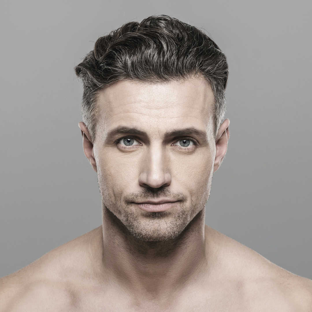 Nahaufnahme eines Mannes in den 30ern ohne Hemd, mit kurzen dunklen Haaren, leichtem Bart und blaugrauen Augen, die direkt in die Kamera blicken.