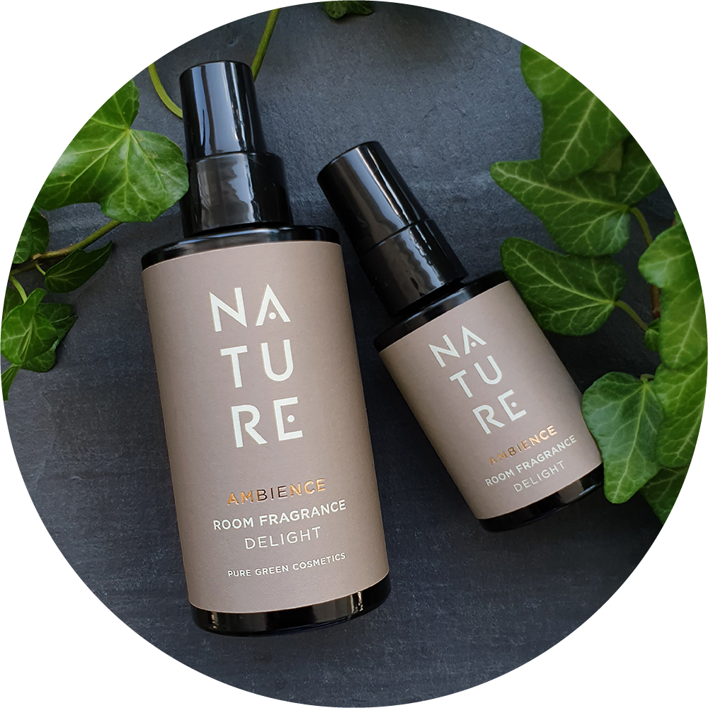 Zwei Sprühflaschen Nature Ambience Raumduft von Pure Green Cosmetics, umrahmt von grünen Efeublättern, auf dunkler Oberfläche arrangiert.