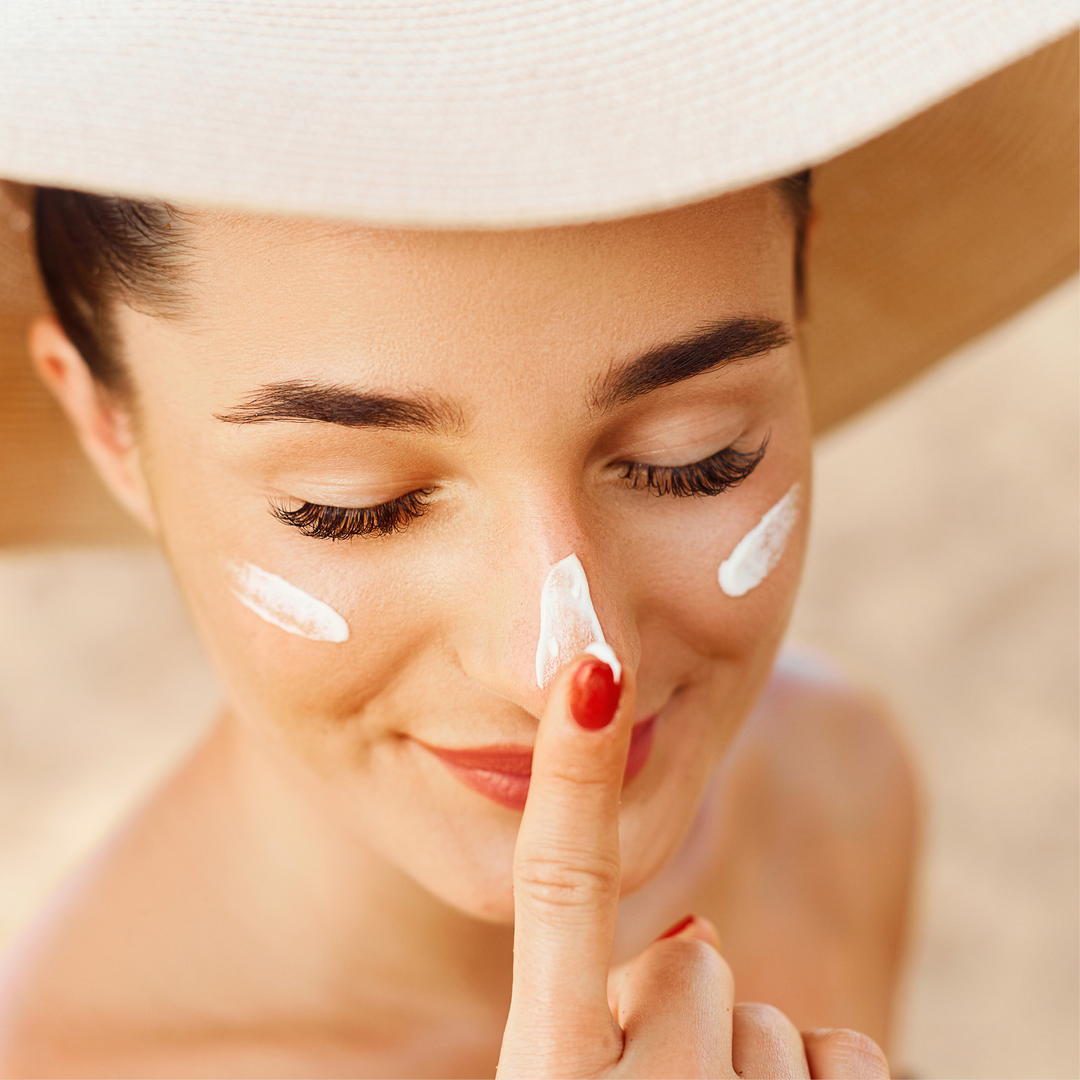 Nahaufnahme einer Frau, die am Strand Sonnencreme auf ihr Gesicht aufträgt. Sie trägt einen Strohhut und lächelt, während sie die Creme verteilt.