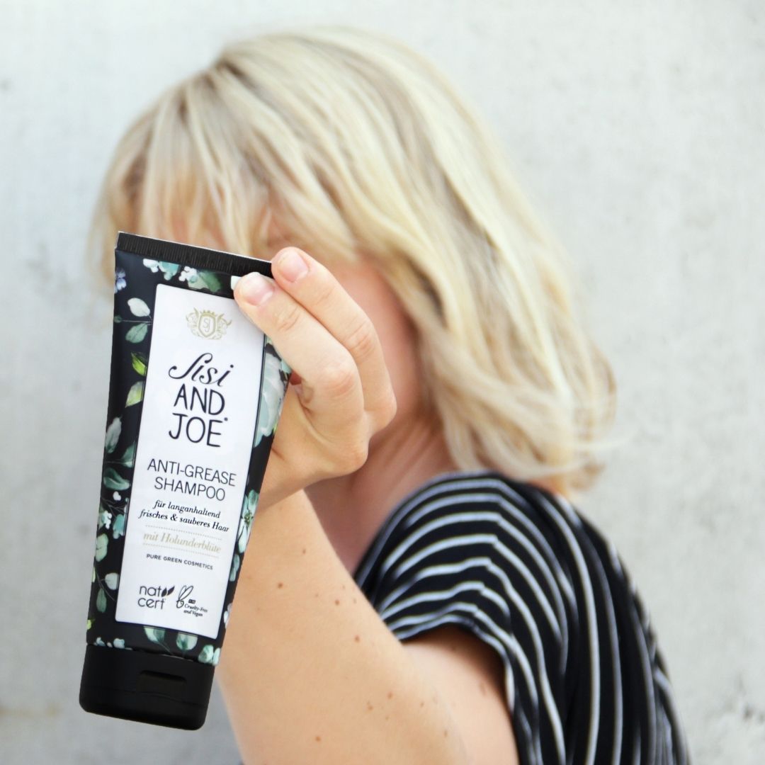 Person hält Sisi and Joe Anti-Fett-Shampoo mit floral verziertem Etikett, zertifiziert von natrue, für gründliche Reinigung und langanhaltende Frische, bio und vegan.