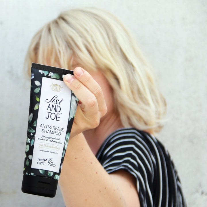 Person hält Sisi and Joe Anti-Fett-Shampoo mit floral verziertem Etikett, zertifiziert von natrue, für gründliche Reinigung und langanhaltende Frische, bio und vegan.