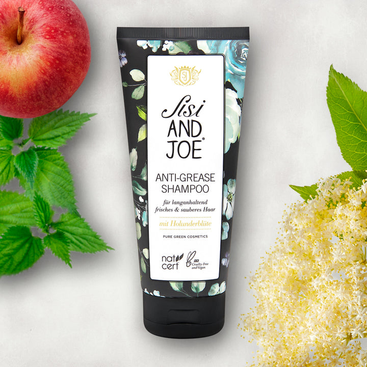 Anti-Fett-Shampoo mit Apfelextrakt & Holunderblütenextrakt in schwarzer Tube, umgeben von Apfel, grünen Blättern und Holunderblüten. Bio, vegan, cruelty-free, zertifiziert.