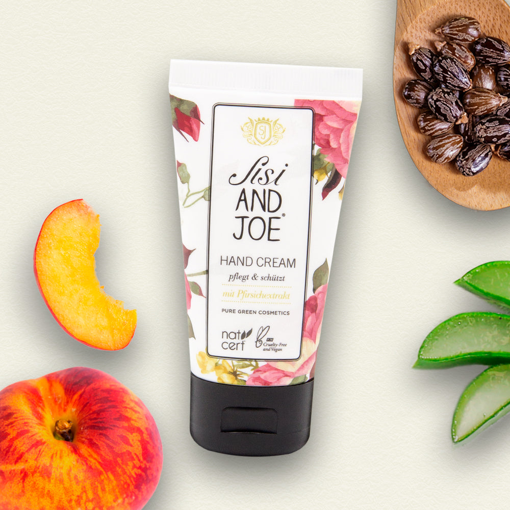 Peachy Kiss Handcreme mit Pfirsichextrakt, Aloe-Vera, umgeben von Pfirsichen, Rizinusöl und Aloe-Blättern. Bio, vegan, pflegt Hände und Nägel seidig-weich.