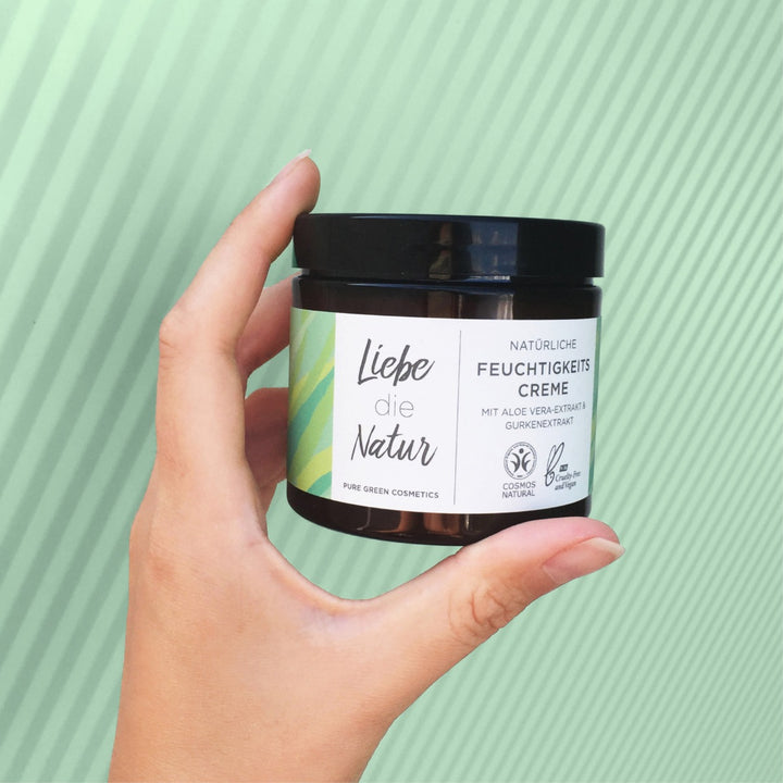 Hand hält einen kleinen, bernsteinfarbenen Kosmetiktiegel mit schwarzem Deckel, beschriftet mit Liebe die Natur Feuchtigkeitscreme mit Aloe-Vera & Gurkenwasser, vegan und COSMOS Natural zertifiziert.