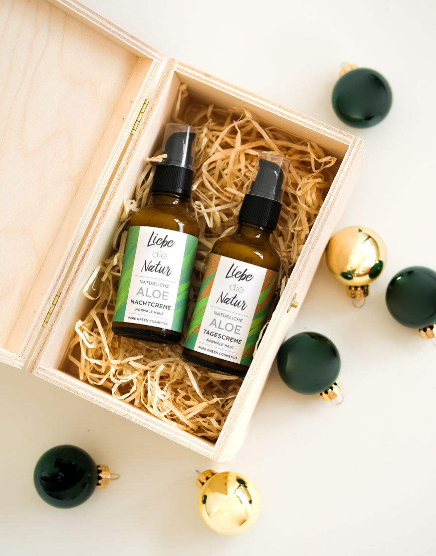 Gesichtspflegeset für wohltuende & entspannende Gesichtspflege | Bio & Vegan: Unvollendete Holzbox mit Aloe Tagescreme und Nachtcreme in Bernsteinflaschen, umgeben von Weihnachtskugeln.