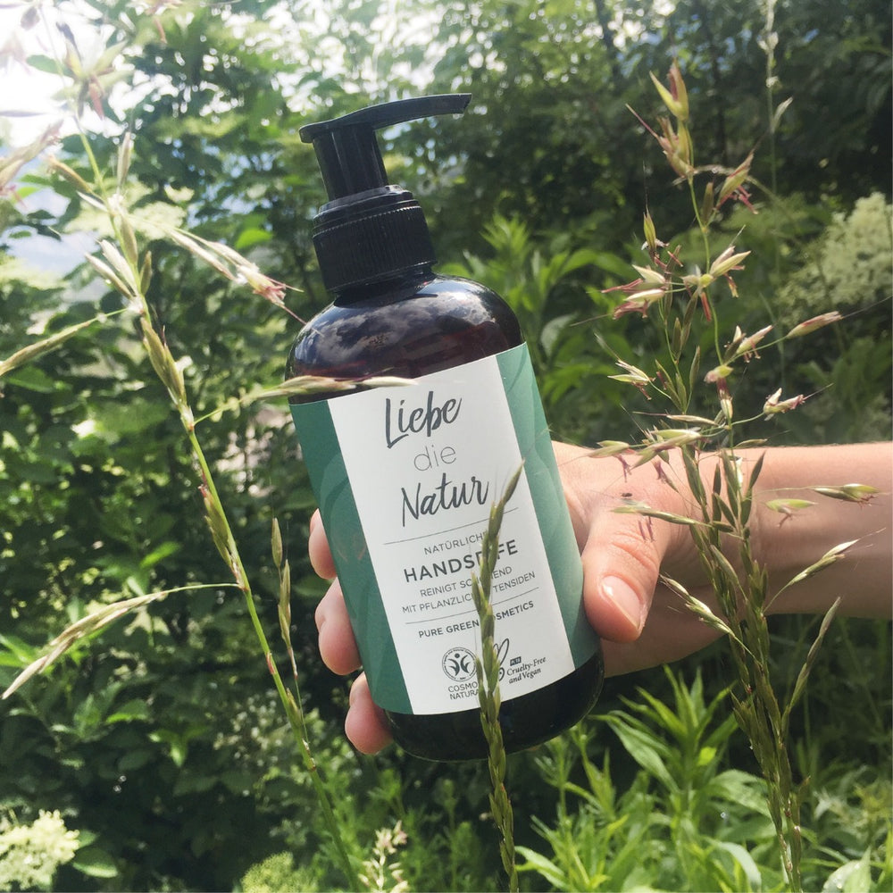Eine Person hält eine Handseife mit pflanzlichen Tensiden & Aloe-Vera-Gel für sanfte Reinigung & zartes Hautgefühl gegen grünen Hintergrund, vegan und COSMOS-zertifiziert.