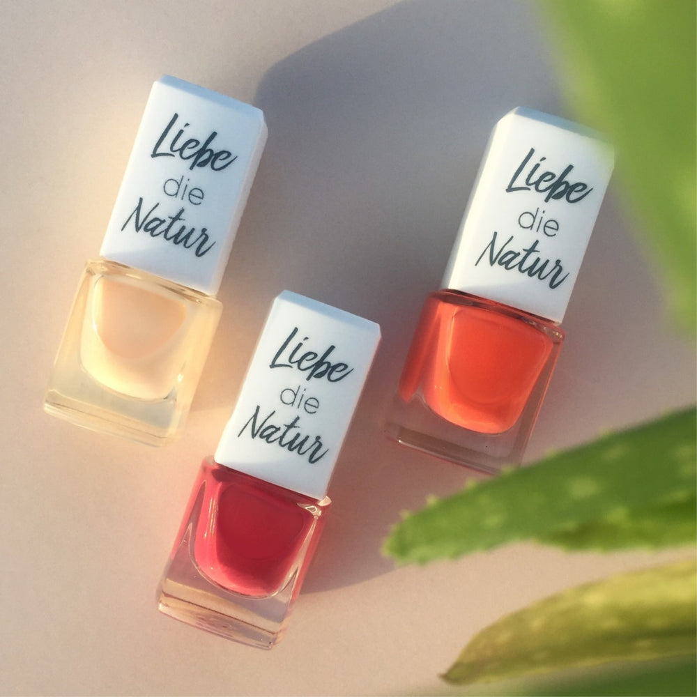 Drei Flaschen Nagellack mit wasserbasierter Formel und natürlichen Farbpigmenten in Nude, Orange und Korallrot auf hellem Untergrund, neben einem grünen Blatt.