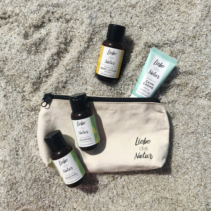 Toiletry-Tasche mit Reisegrößen von Liebe die Natur: Shampoo mit Zitronenmelissenextrakt & Brennnesselextrakt, Duschgel, Body Lotion, Zahnpasta auf Sand.