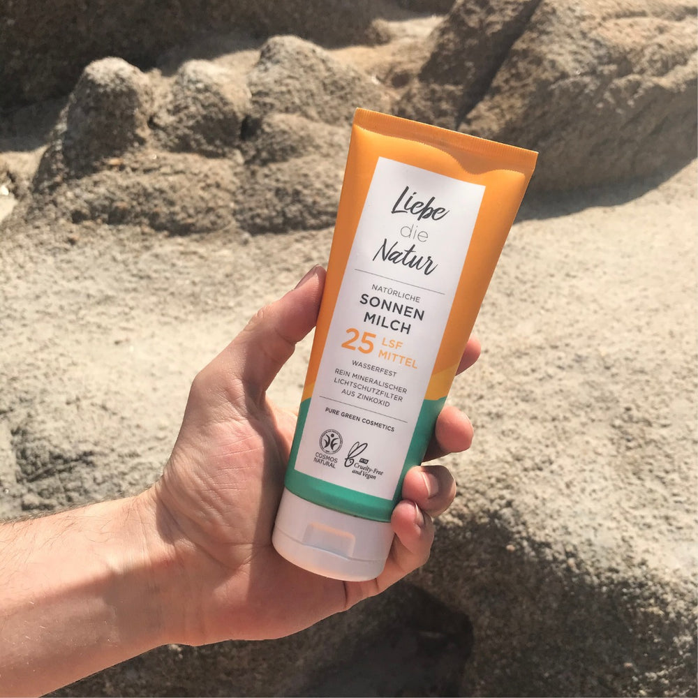 Hand hält eine Sonnencreme LSF 25 mit Zinkoxid und Sonnenblumenöl von Liebe die Natur auf einem Felsenstrand, bietet natürlichen, wasserfesten Sonnenschutz.