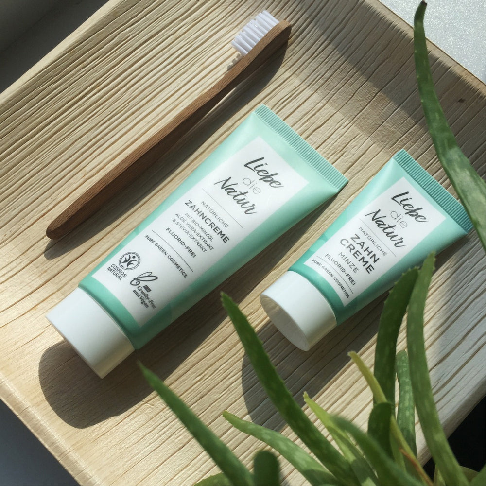 Bambuszahnbürste und zwei Tuben Liebe die Natur Zahncreme mit Bio-Pfefferminzöl auf Holztablett, begleitet von Aloe Vera für pflanzliche Frische.