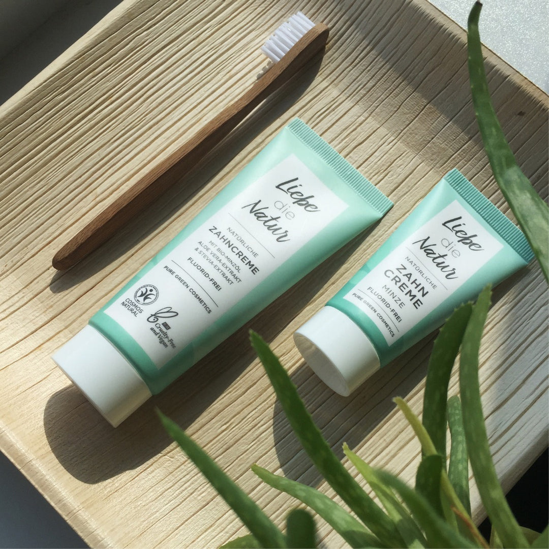 Bambuszahnbürste und zwei Tuben Liebe die Natur Zahncreme mit Bio-Pfefferminzöl auf Holztablett, begleitet von Aloe Vera für pflanzliche Frische.