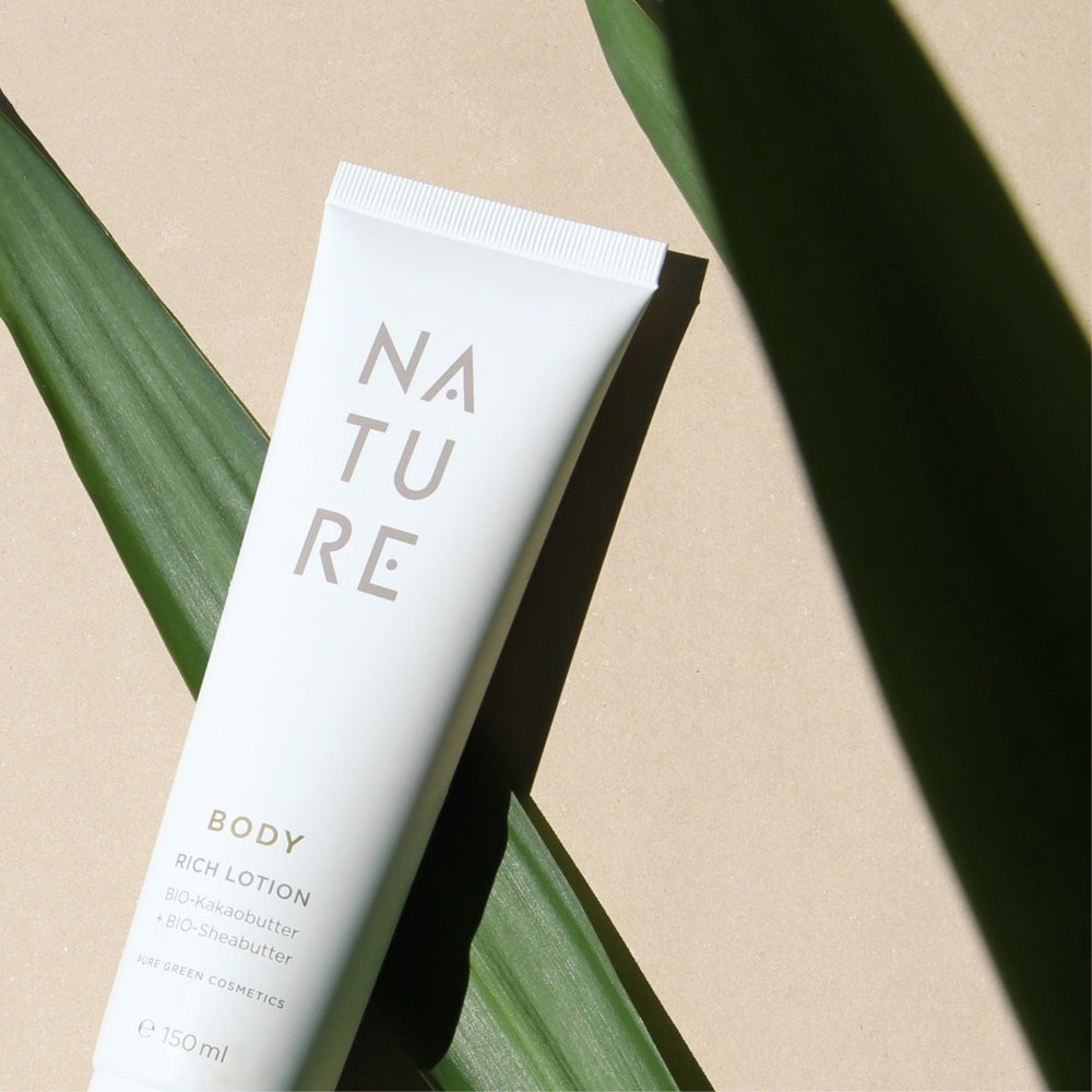 Weiße Tube Bodylotion mit NATURE Aufschrift, enthält Bio-Kakaobutter und Aprikosenkernöl. Platziert auf beigem Hintergrund mit grünen Blättern für einen natürlichen Look.
