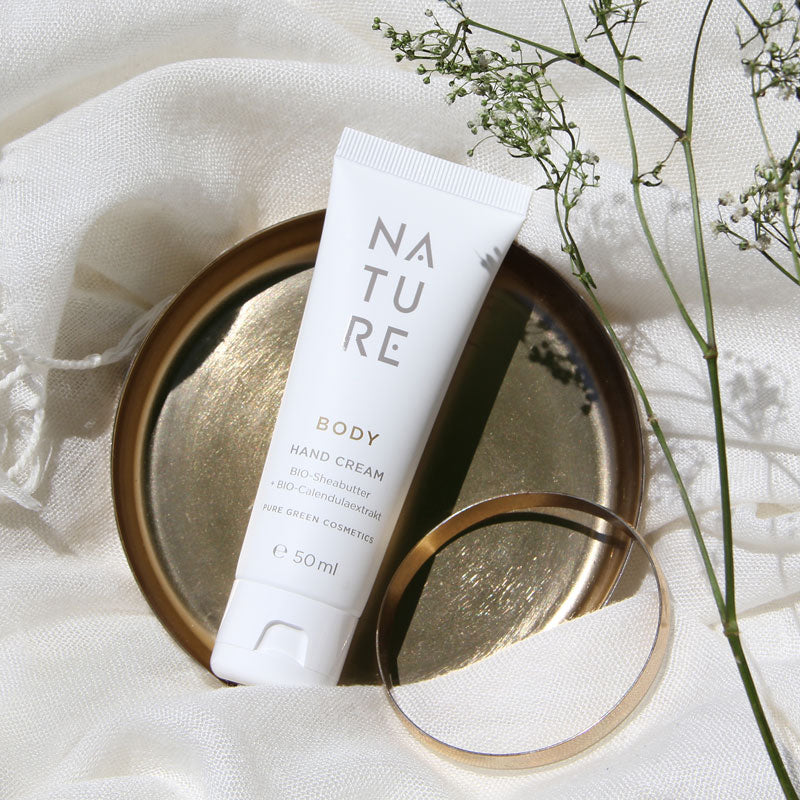 Handcreme mit Sheabutter & Calendula auf goldener Schale, umgeben von einem goldenen Armreif und weißen Blumen. Intensive Feuchtigkeit, bio & vegan.