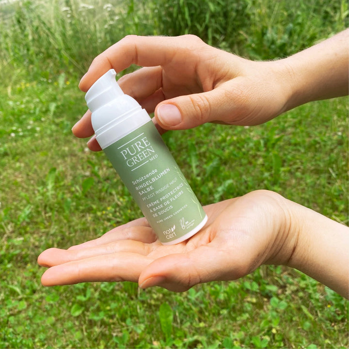 Hände halten eine Pumpflasche Pflegende Ringelblumensalbe mit BIO-Nachtkerzenöl & Sheabutter, eine schützende Salbe für trockene Haut, im Freien.