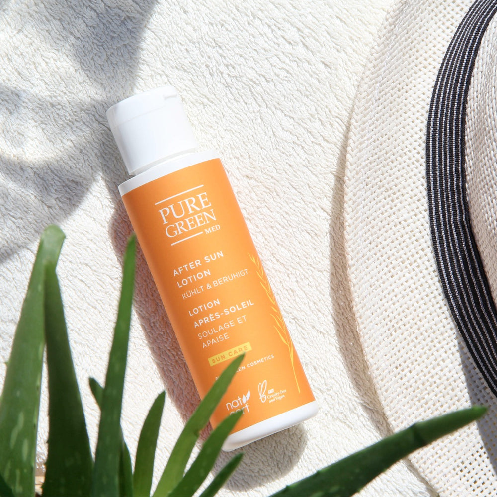 Flasche Pure Green Med After-Sun-Lotion mit Aloe-Vera und Gurkenwasser auf Handtuch, daneben Aloe-Blätter und Sonnenhut. Ideal für regenerierte, gekühlte, sonnenstrapazierte Haut; bio und vegan.